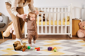 young mother holding hands of little son walking near toys and crib in nursery room, support - 731972684