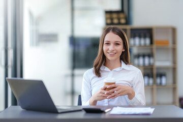 Smiling young businesswoman holding a coffee cup, sitting confidently in a modern office with a...