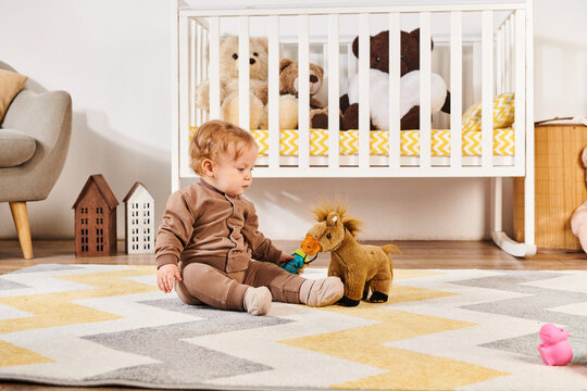 adorable toddler boy sitting on floor and playing with toy horse near crib in nursery room