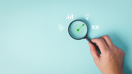 A hand holding a magnifying glass focuses on a target symbol, surrounded by icons representing analytics, ideas, and financial growth on a light blue background.