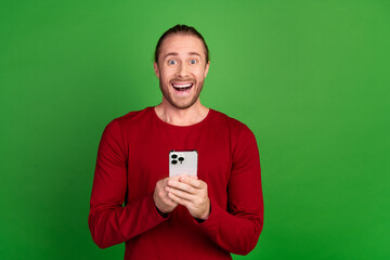 Portrait of satisfied impressed guy with long hair wear red shirt hold smartphone asstonished staring isolated on green color background