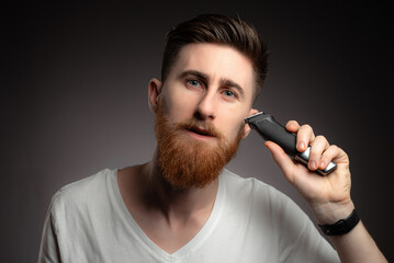 Portrait of handsome man shaving his beard with electric shaver in morning, against grey background