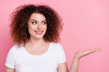 Portrait of cheerful girl with wavy hairdo wear white t-shirt look at product on arm empty space isolated on pink color background