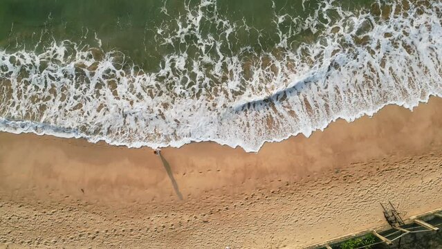 Aerial view captures woman strolling along seaside. Beach walk in slow motion, tranquil shore waves wash over sandy beach. Sunlit holiday, coastal travel, relaxing oceanside journey.