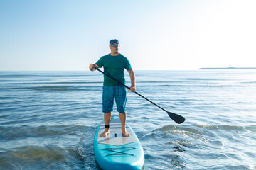 A middle-aged man in shorts and a T-shirt stands on a SUP board with a paddle near the sea.