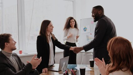 Boss shaking hand of young shy woman congratulating successful employee with promotion, hiring intern, appreciating for good work result, rewarding while business team applauding supporting colleague