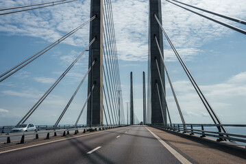 A clearskied modern suspension bridge with twin towers and cables, calm seas on either side. An empty lane, white car seen, possibly the Oresund Bridge linking Copenhagen to Malmo.