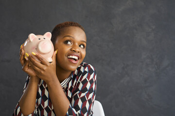 Happy dark skinned woman listens to the sound of coins in a piggy bank and dreams. Close up of...