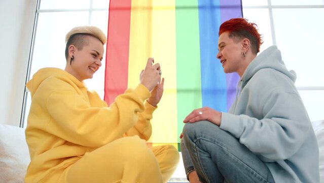In a serene domestic setting, one woman lovingly captures her partner's image on her phone as they sit by a large window draped with the iconic LGBT flag, their home a haven of pride and love.