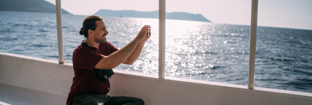 A young, contented man, a tourist rides a boat to the sea, takes pictures on the phone, enjoys a sunny day.