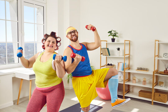 Funny Overweight Happy Fat Woman With Bearded Man In Bright Sportswear Looking At The Camera And Doing Fit Exercises With Dumbbells At Home Together. Concept Of Couple Family Sport And Fitness.