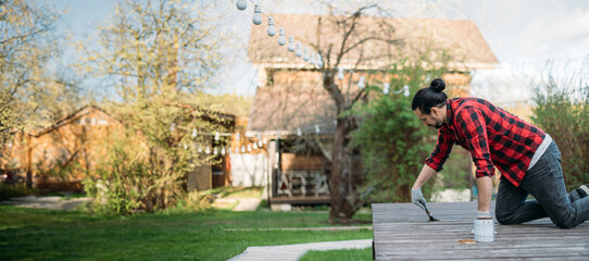 A young man in home clothes paints a wooden deck in the garden with a brush.