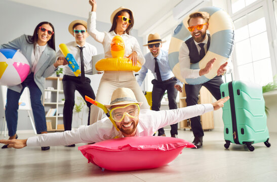 Funny business team ready for summer holiday together. Man in swimming mask and sun hat on water mattress on office floor, friends having fun with lifebuoy, beach ball, toy gun, luggage in background