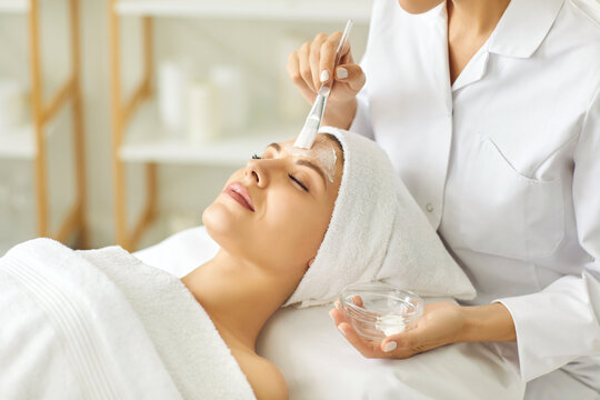 Woman customer enjoying professional skincare at spa salon. Cosmetologist with brush and glass bowl applies moisturizing beauty mask on face of relaxed young woman client. Facial treatment concept