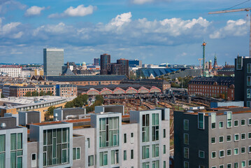 Overlooking a cityscape, modern homes with sleek designs beside a classic redroofed structure, likely in Copenhagen. Partly cloudy skies and active construction suggest expansion.