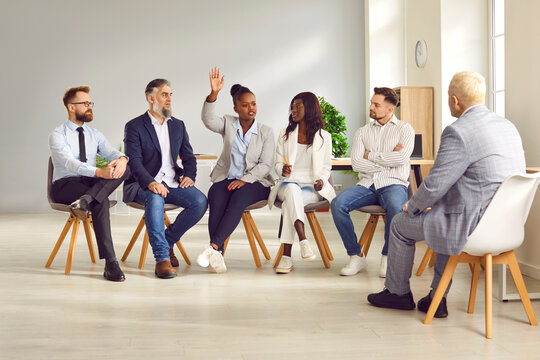 Portrait of african american woman raising hand to ask or answer a question on a meeting. Group of company employees or a team of staff sitting on chairs in a row and listening to their leader.
