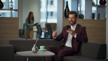 Joyful businessman drinking coffee holding smartphone in office. Manager relax