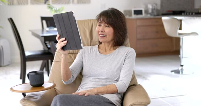 Smiling woman happy enjoying video chat talking using mobile tablet technology sitting in comfortable home leather chair.