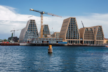 Modern waterfront construction in Copenhagen with saillike buildings and cranes against a partly cloudy sky, reflecting on calm waters, indicating a harbor redevelopment.