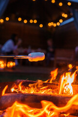 A closeup view of a marshmallow toasting over a fire pit, turning slightly brown, with folks and gentle lighting in the backdrop, possibly in Copenhagen. - obrazy, fototapety, plakaty