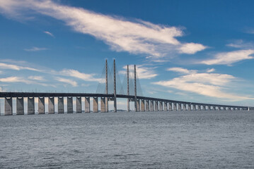 The Oresund Bridge, connecting Copenhagen, Denmark, with Malmo, Sweden, arches over calm waters, its high pylons and cables creating a striking silhouette against a clouddotted sky.