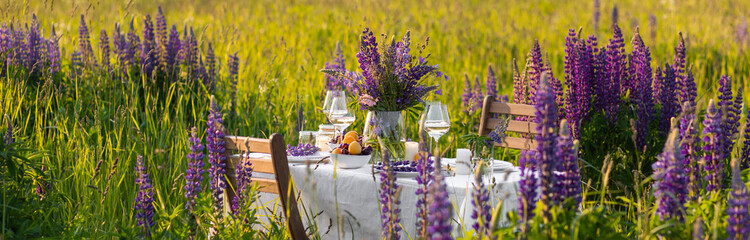 Elegant gorgeous wedding table decor or romantic dinner arrangement outdoors in blooming field. Purple lupine flowers, candles, fruits and wine, wooden furniture. Sunset, summer, golden hour banner - obrazy, fototapety, plakaty
