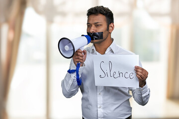 Indian man with taped mouth holding poster with inscription Silence. Censored male protester with...