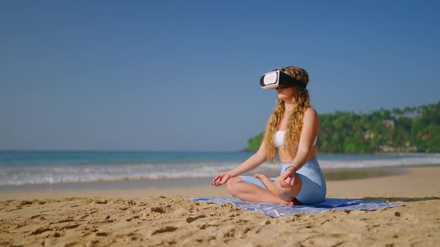 Woman in VR headset sits in lotus pose on beach, meditates in virtual reality. Mindful tech user finds tranquility, immerses in digital mindfulness retreat. Future of calm interactive experiences.