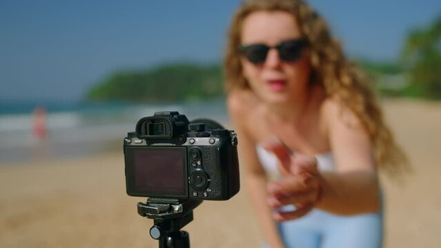 Woman in tube top and jeans films dance routine on beach with camera on tripod. Content creator performs, records trendy moves for followers. Sunny day, seascape backdrop, social media focus.