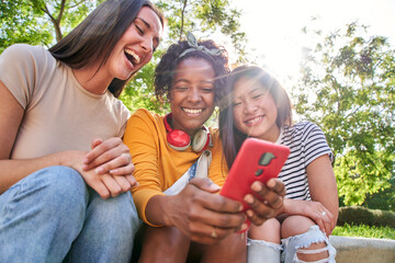 Group joyful friends using mobile sitting outdoor. Three laughing multiracial women viewing social...
