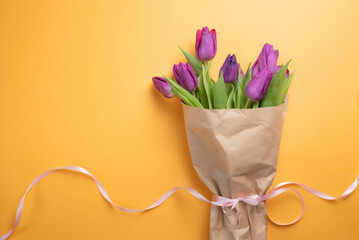 Bouquet of fresh, purple tulip flowers wrapped in brown paper, tied with a long, pink ribbon, on a yellow background.