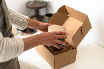 Artisan placing a ceramic vase into a cardboard box with protective packing material for safe...