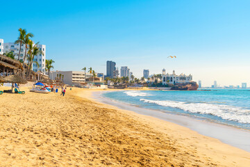 The wide sandy Playa Gaviota beach full of resorts and restaurants, with Valentinos Disco and the skyline of Mazatlan, Mexico behind, on a sunny day along the Golden Zone. - obrazy, fototapety, plakaty