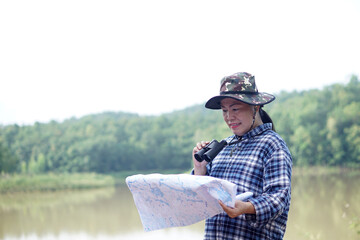 Obraz premium Asian woman exploror holds binocular and looking at paper map to explore nature at the lake area. Concept, nature exploration. Ecology study. Pastime activity, lifestyle. Explore environment 