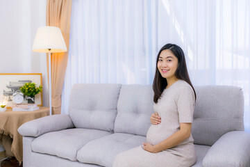 Smiling beautiful pregnancy woman sitting on sofa  by the window of her home, showcasing beauty and a positive lifestyle.Asian pregnant female.White background with copy space.Hand on tummy or belly.