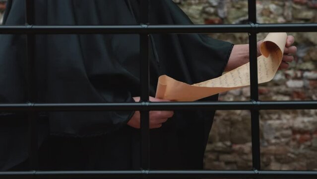 A monk in a black cassock unrolls a scroll and reads behind a grille against a stone wall.