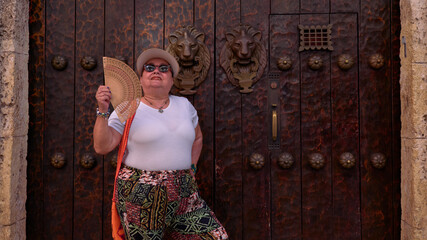 A medium shot portrait of an older woman dressed in colorful pants and a white blouse, wearing a visor and dark sunglasses, cooling herself with a fan. In the background, there's a Colonial door