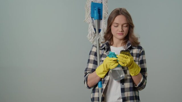 A Happy Young Woman in Yellow Gloves with a Mop, Spray Bottle and a Smartphone, Rejoices at Success on the Phone on the Gray Background.Virtual Consultations for Appliance Cleaning.