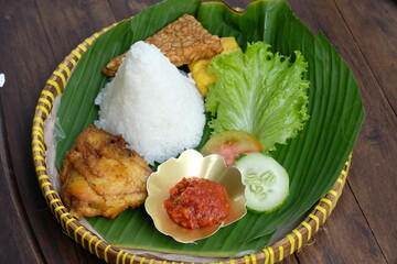 Ayam Goreng Tempe Nasi Tumpeng is indonesian traditional fried chicken. Served with fried tofu, fried tempeh, sambal (chili sauce), sliced cucumber , sliced tomato, and fresh lettuce. Banana leaves. 