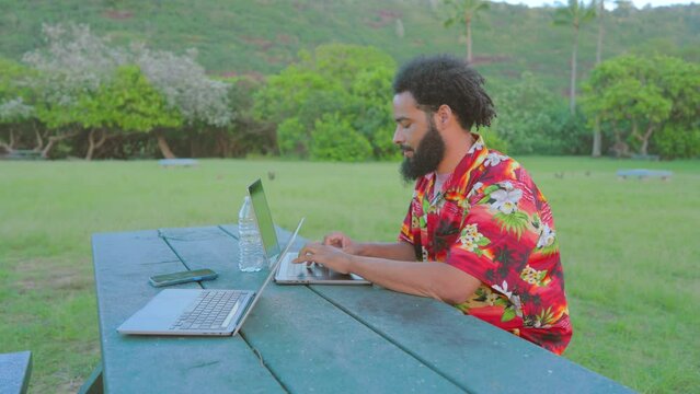 Remoter working black man leisurely sits at an outdoor table working on a laptop computer with his friends laptop surrounded by lush grass and plants.