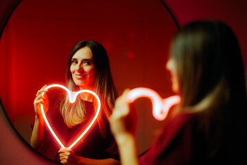 Happy Woman Holding a Neon Heart Looking in the Mirror. Cheerful self-loving person accepting...