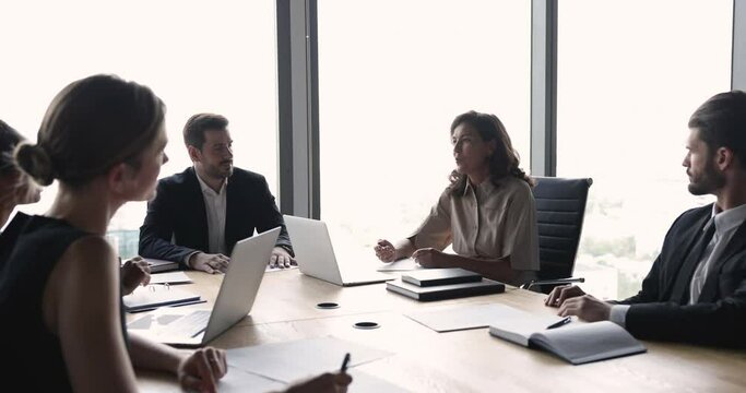 Mature project manager woman speaking at meeting with business colleagues, sitting at large table, talking to coworkers, offering ideas for brainstorming, cooperation, teamwork strategy