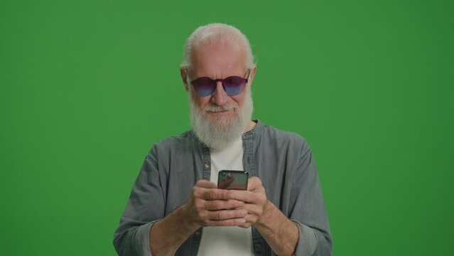 Green Screen.A Smiling Old Man with Gray Beard and Funny Glasses Scrolls on the Phone. Happy Elderly Man Shops Online.Digital Literacy and Tech Training for Seniors.