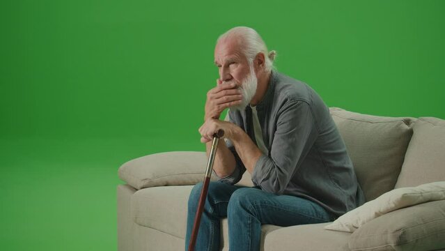 Green Screen. A Sad Old Man with a Stick Is Sitting On A Sofa. An Elderly Man Leaning His Head On A Stick and Rubs His Gray Beard. Community and Social Support for Seniors.