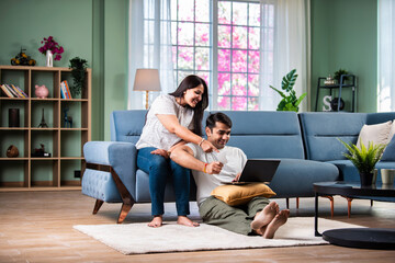 Young and happy Indian couple sitting on sofa at home, using a laptop for shopping or video call.
