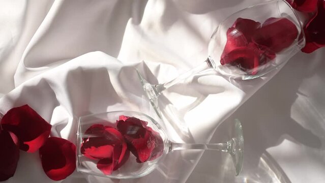 Two wine glasses with rose petals on white bed sheet in sun