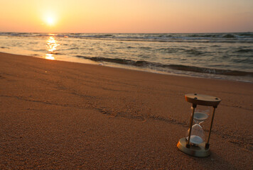 Fototapeta na wymiar An hourglass (sand clock) lies on the beach sand with the background of the sea and the setting sun with a beautiful orange twilight glow, copy space.