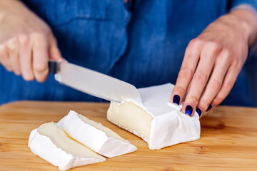 Obraz na płótnie Canvas girl with cheese. female hands with blue nails cutting camembert cheese with a knife on a light wooden kitchen board, cooking concept