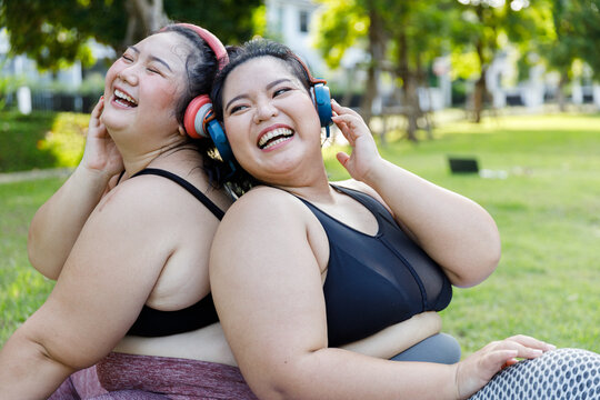 Cheerful laugh smiling friends young asian woman happy wear headphone listen to music in sportswear walking after exercise. Two sporty women body curvy positive having fun workout in garden.