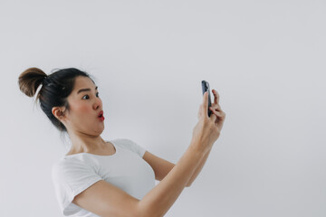 Asian Thai woman holding mobile phone with funny surprised and shocked face, while looking cellphone, standing isolated over white background wall.
